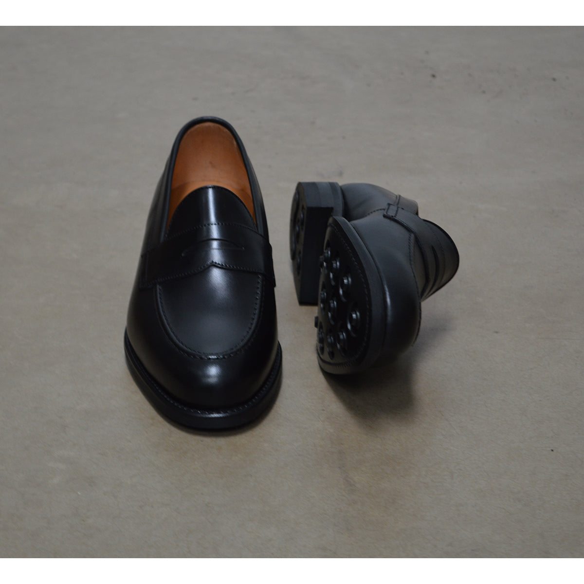 A black calf leather penny loafer with a Dainite rubber sole, showing a classic round toe shape and the iconic penny slot detail.