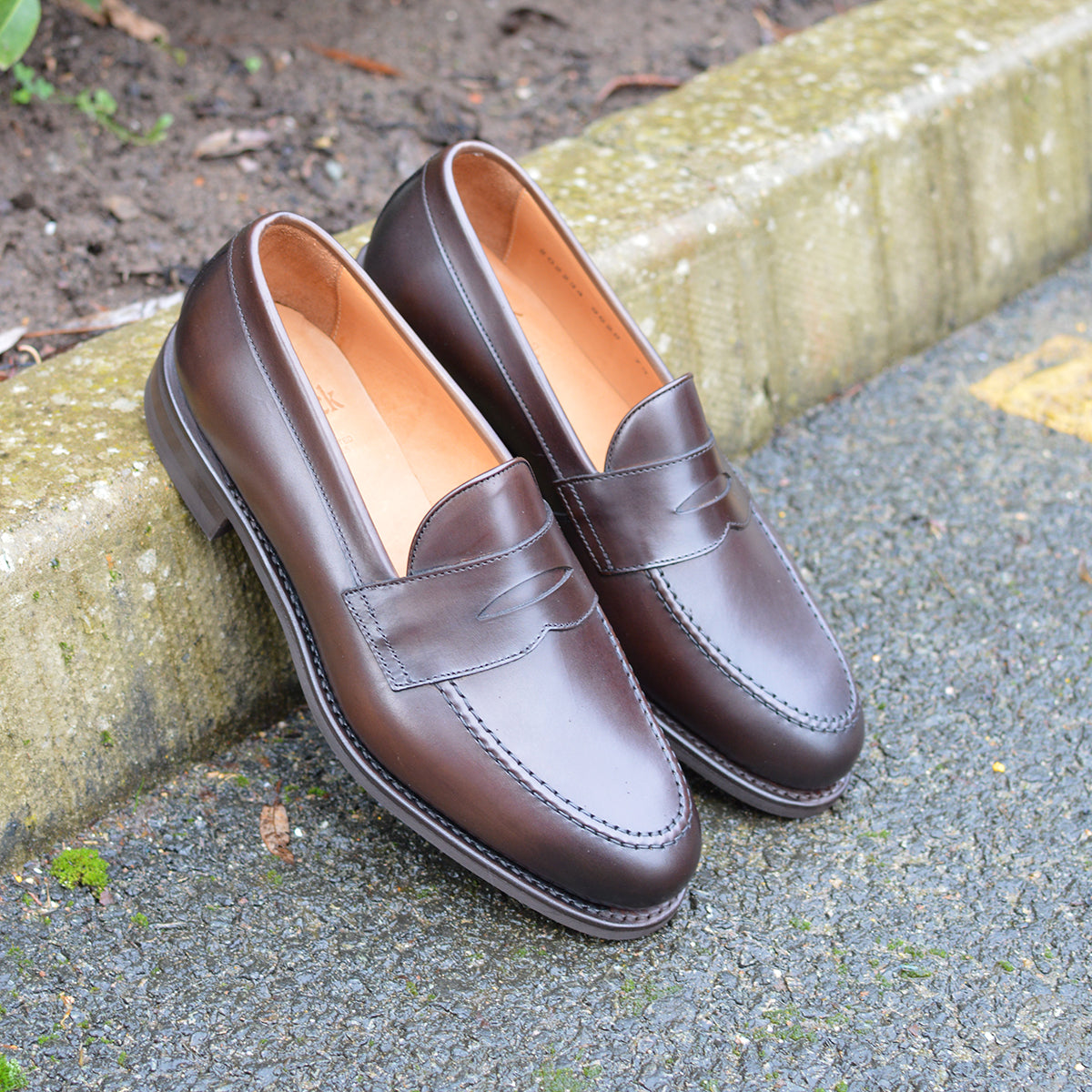 Pair of brown leather loafers on a concrete surface with a stone wall in the background.