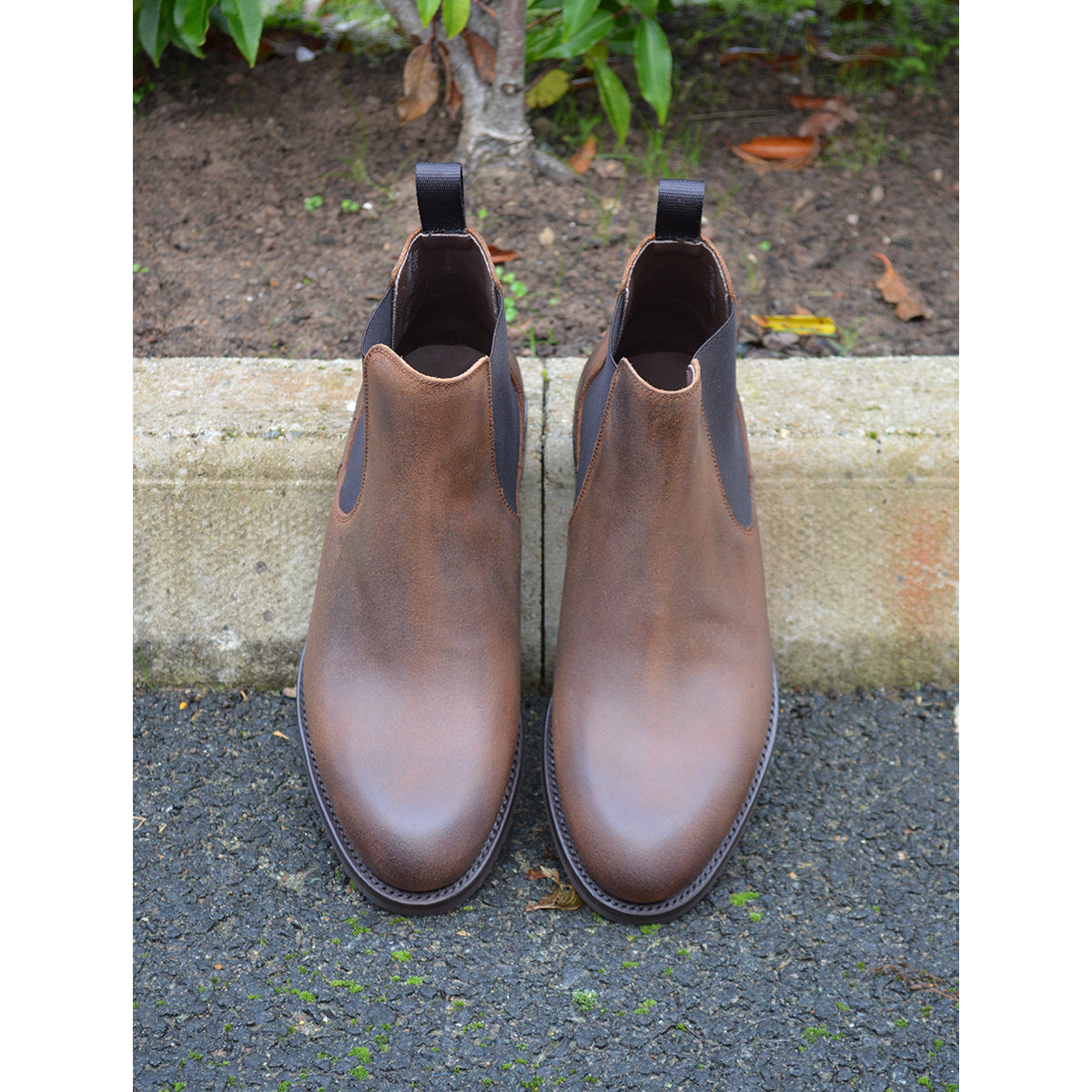 A dark brown Chelsea boot with a waxy leather finish and a contrasting black elastic side panel. The boot has a low heel and a small pull tab at the rear.