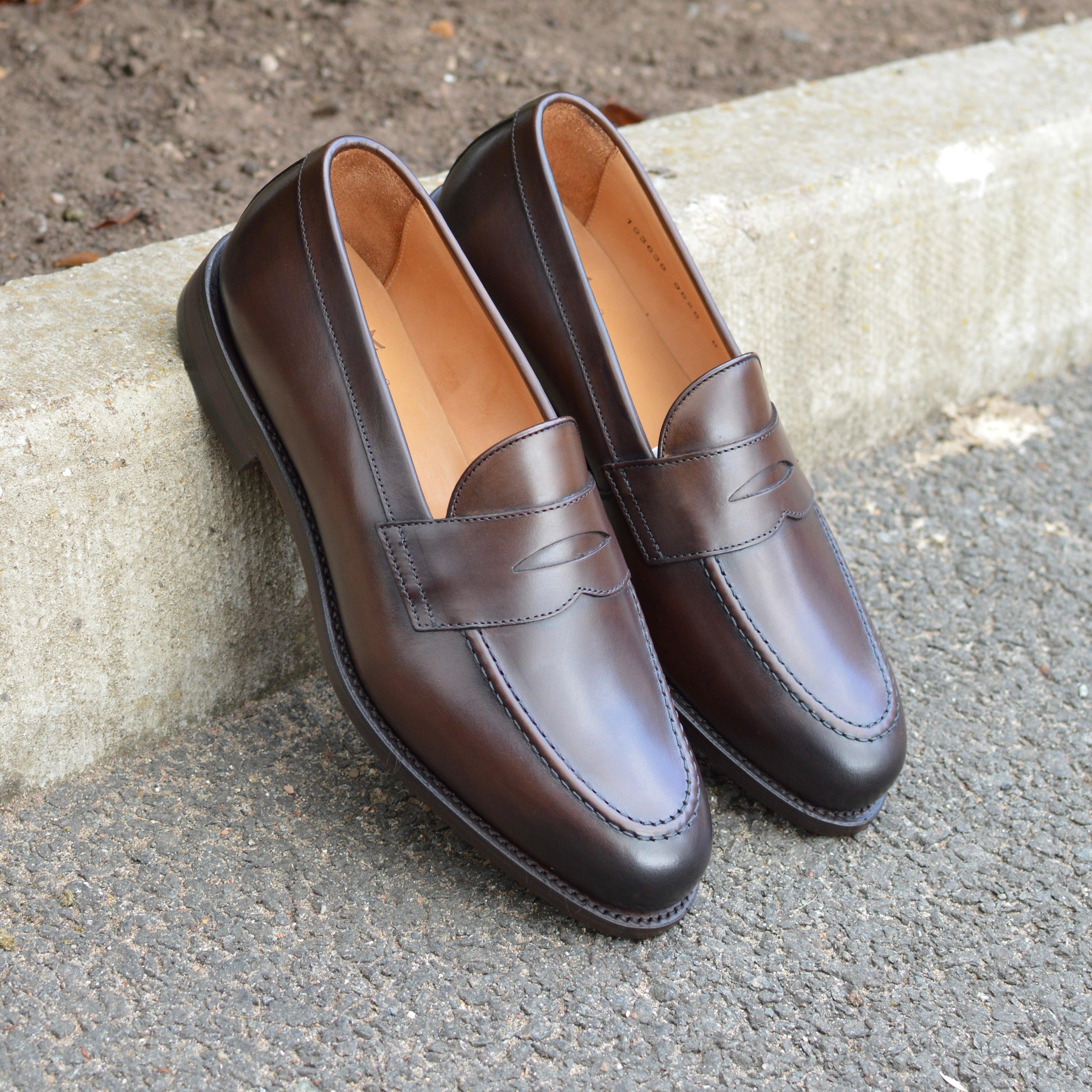 A dark brown Berwick 1707 Penny Loafer with a closed toe and loafers style heel, displayed on a concrete curb background.