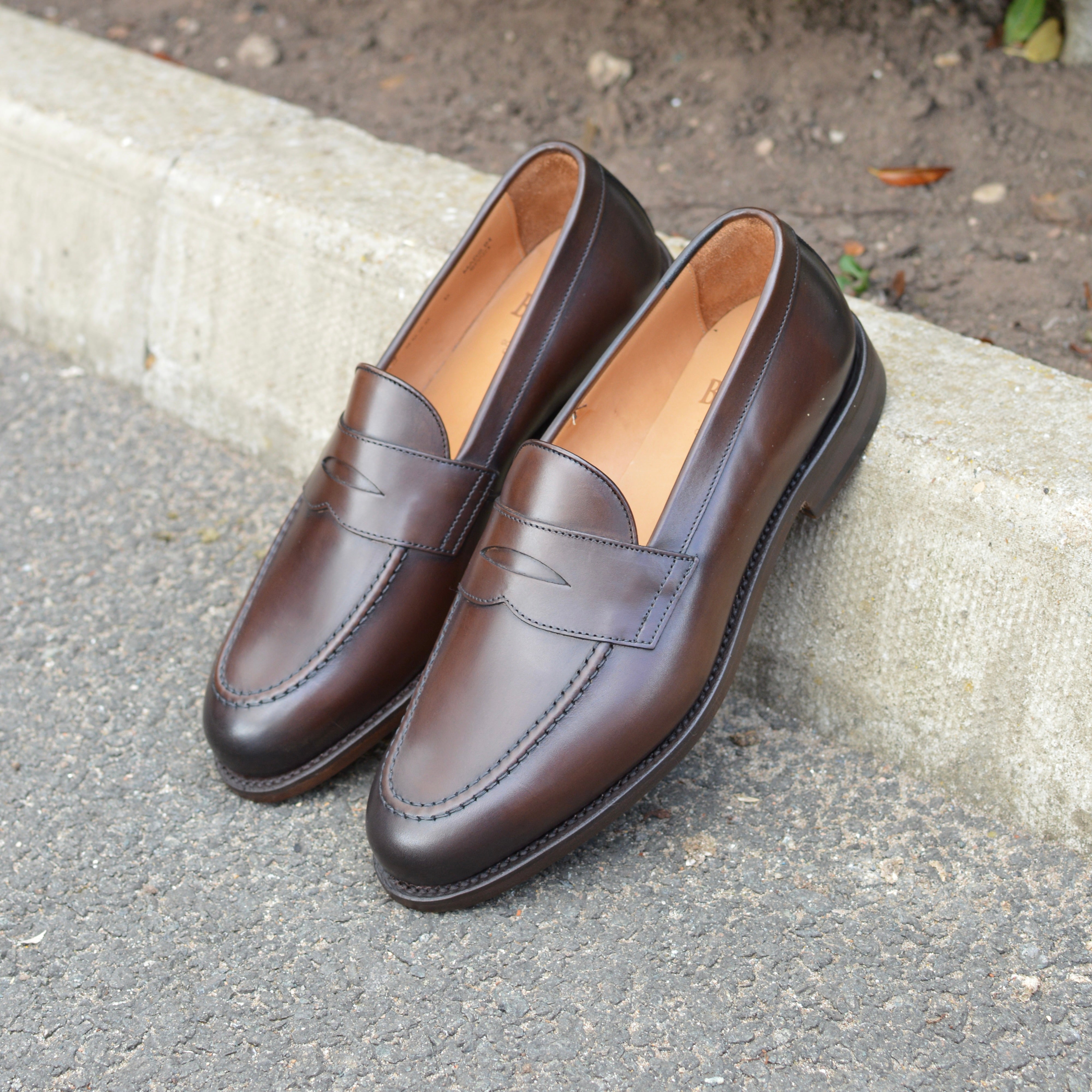 A dark brown Berwick 1707 Penny Loafer with a closed toe and loafers style heel, displayed on a concrete curb background.