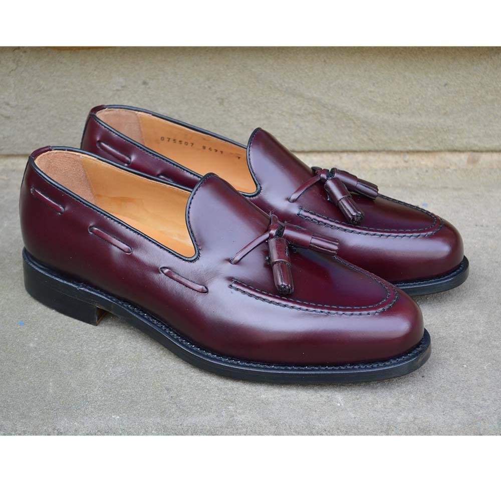 A Berwick 1707 burgundy hi shine leather slip-on shoe with a tassel detail on the vamp, set against a sandstone background.