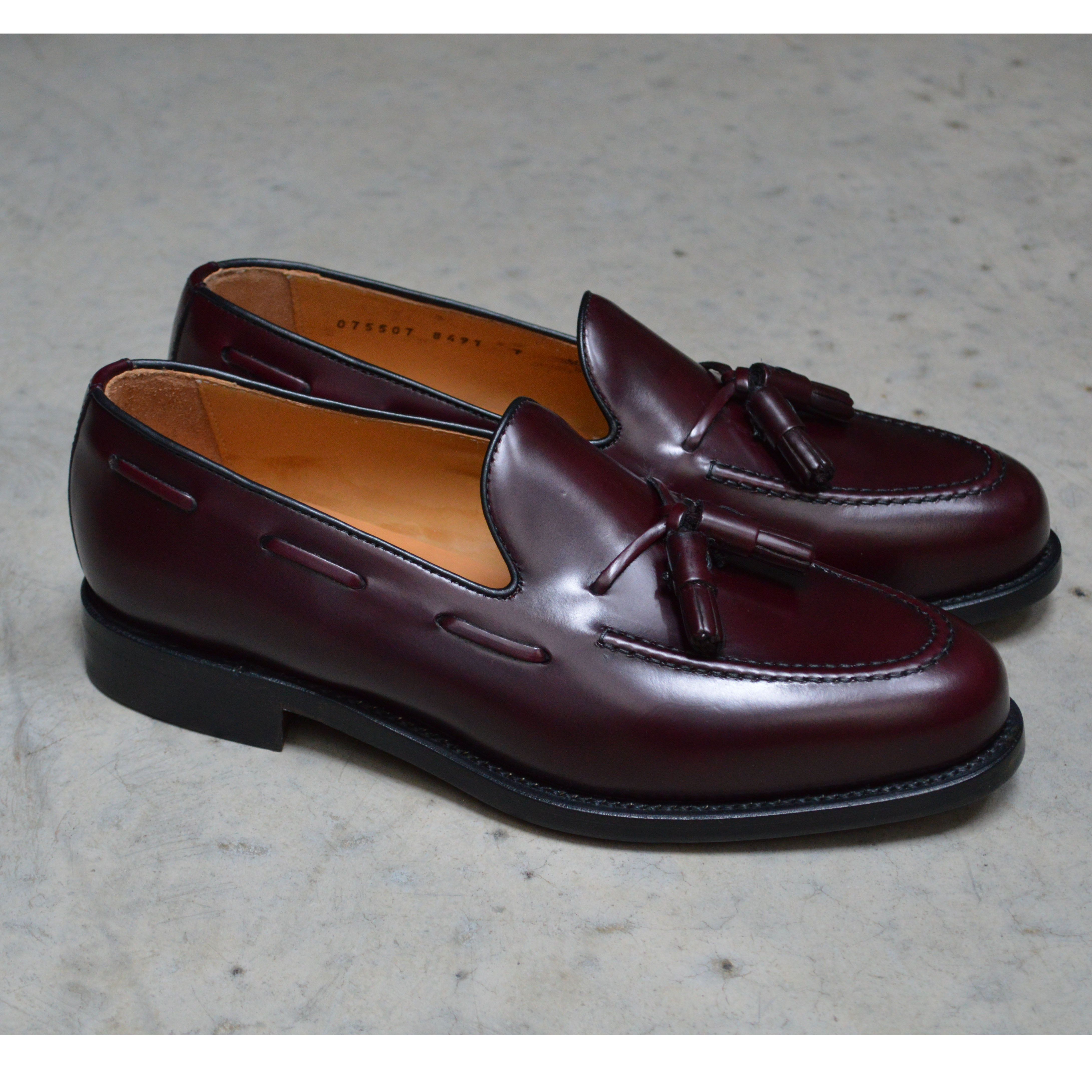 A Berwick 1707 burgundy hi shine leather slip-on shoe with a tassel detail on the vamp, set against a polished concrete background.