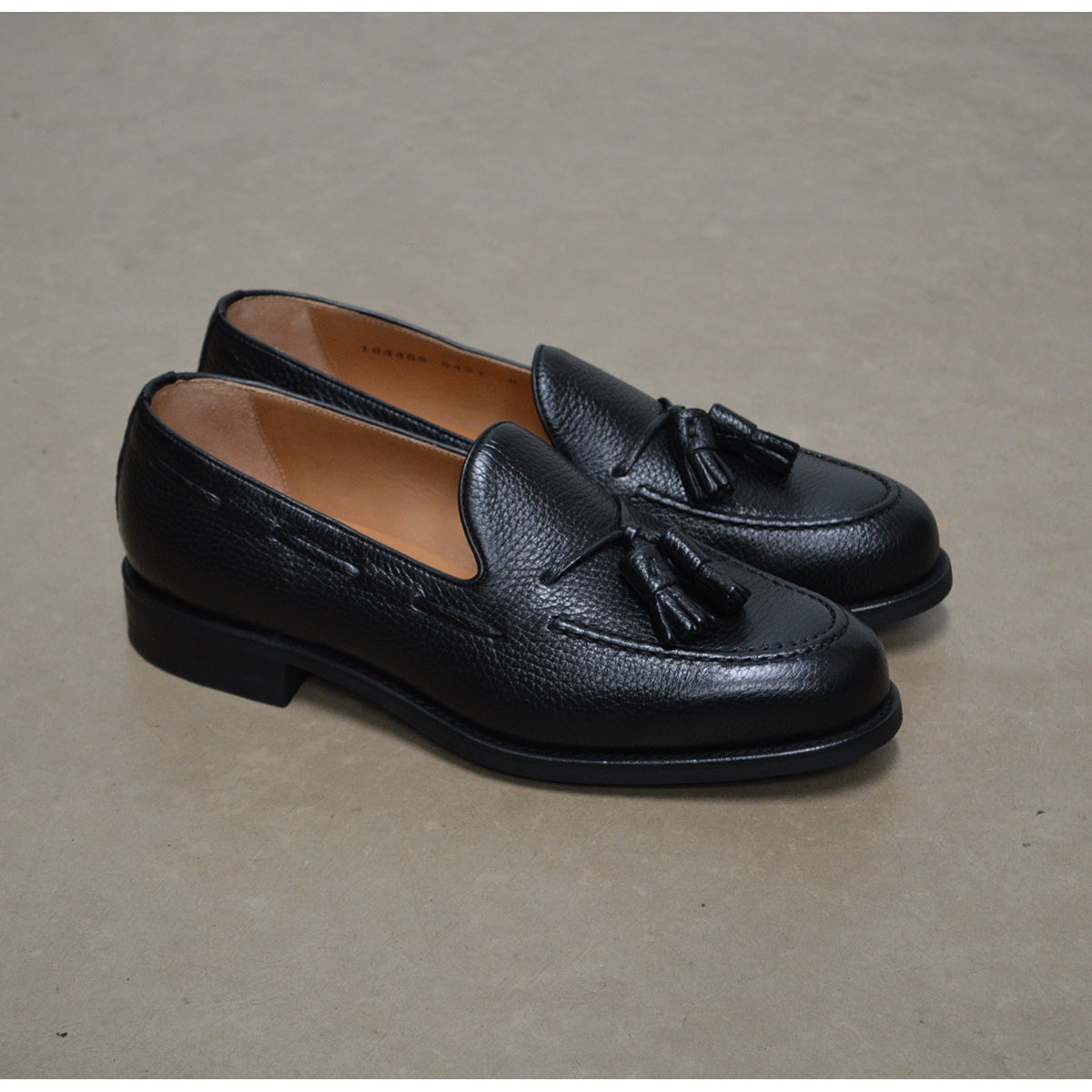 A black deerskin slip-on loafer with a tassel detail on the vamp, featuring a classic round toe and a low heel.