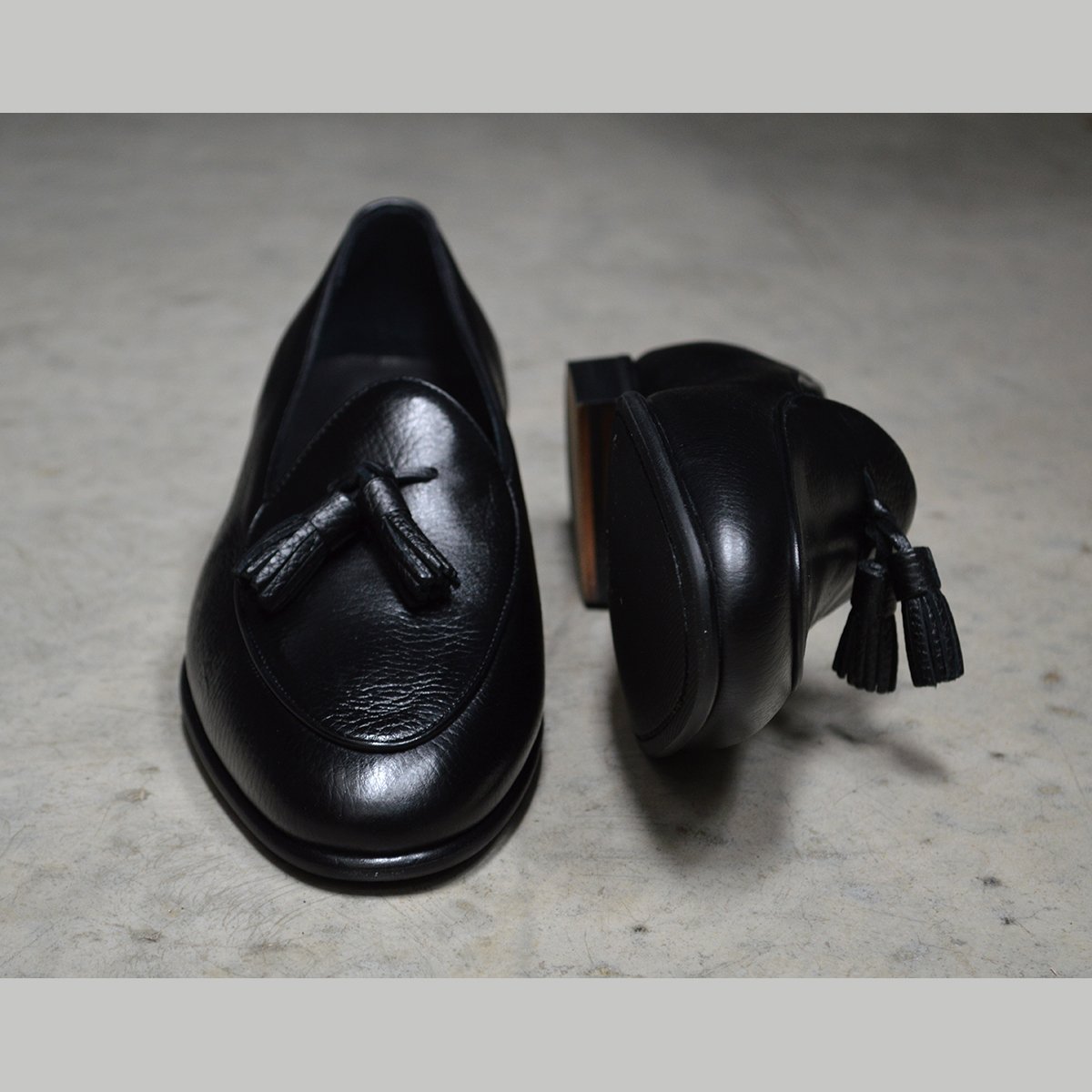 A black deerskin leather tassel Belgian loafer with an almond-shaped toe and a subtle tread sole, displayed against a polished concrete background.