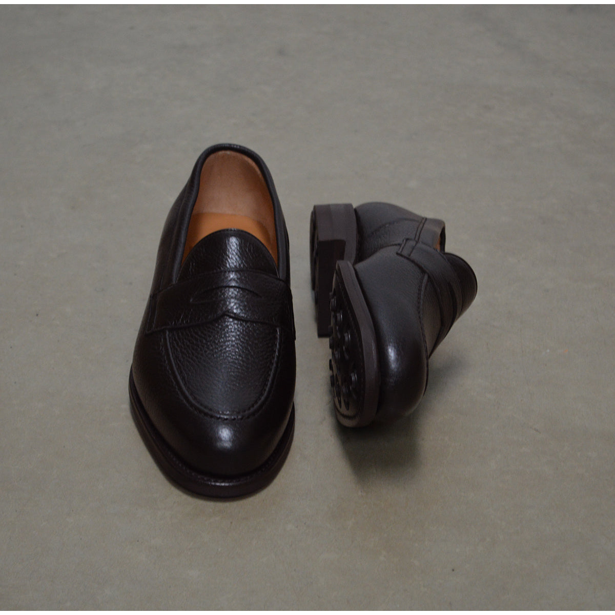 A dark brown deerskin penny loafer with a velvety soft texture and a classic round toe shape on a polished concrete background with Dainite Studded Rubber Sole.