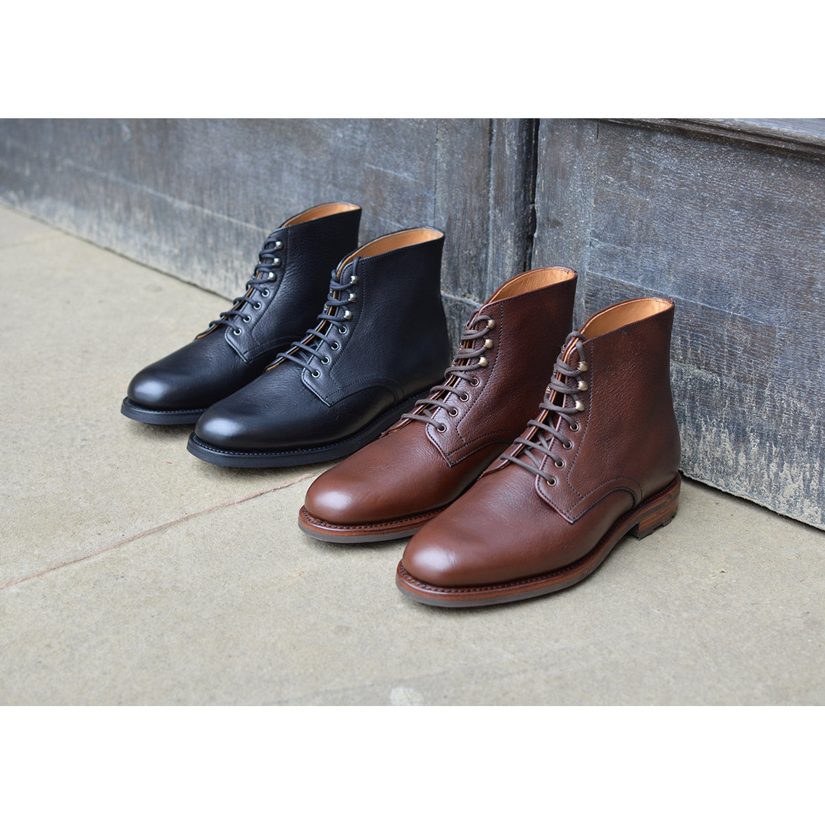Pair of Berwick 1707 black and brown leather boots on a sandstone floor with a wooden rustic door background.