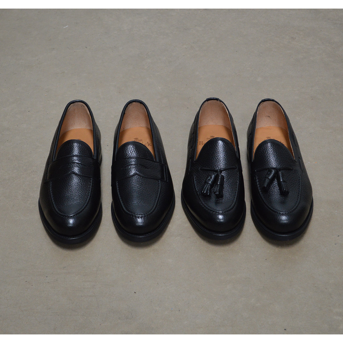 A black deerskin Penny loafer with a velvety soft texture and a classic round toe & Dainite sole, set against a polished concrete floor background.