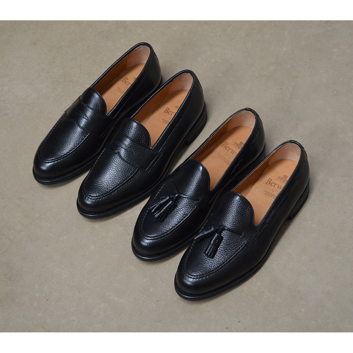 A black deerskin Penny loafer with a velvety soft texture and a classic round toe & Dainite sole, set against a polished concrete floor background.