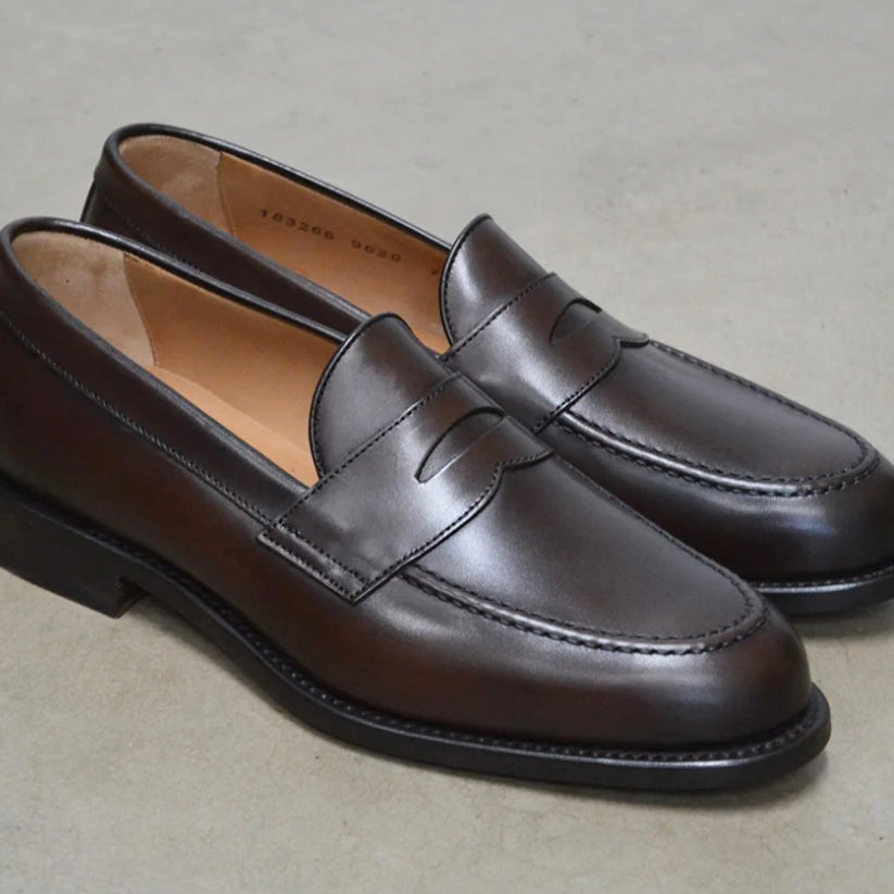 A dark brown Berwick 1707 Penny Loafer with a closed toe and loafers style heel, displayed on a polished concrete background.