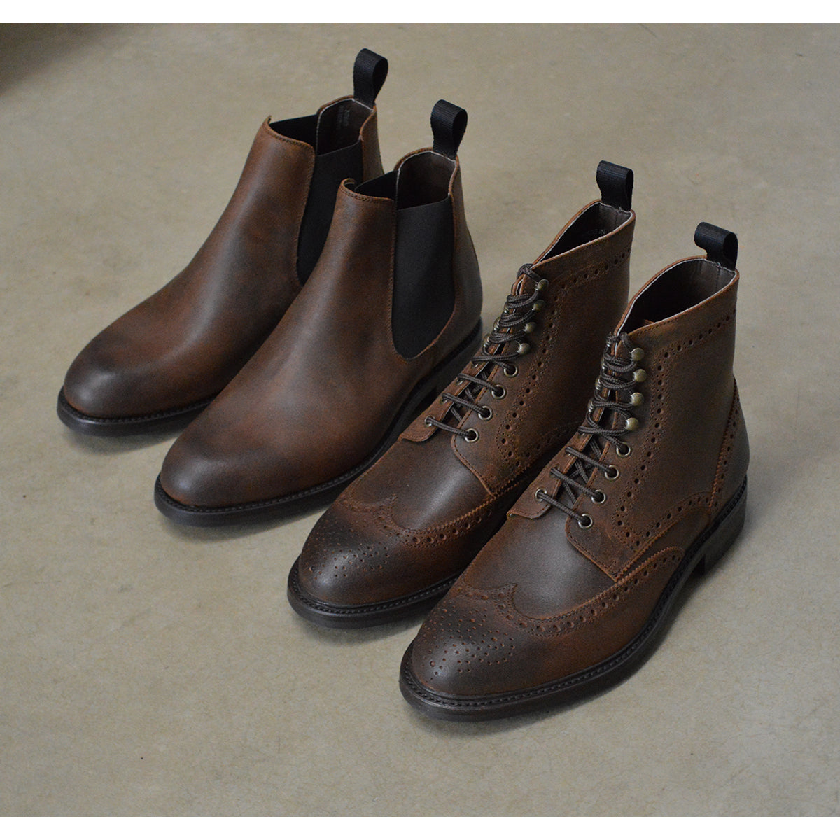 A dark brown Chelsea boot with a waxy leather finish and a contrasting black elastic side panel. The boot has a low heel and a small pull tab at the rear.