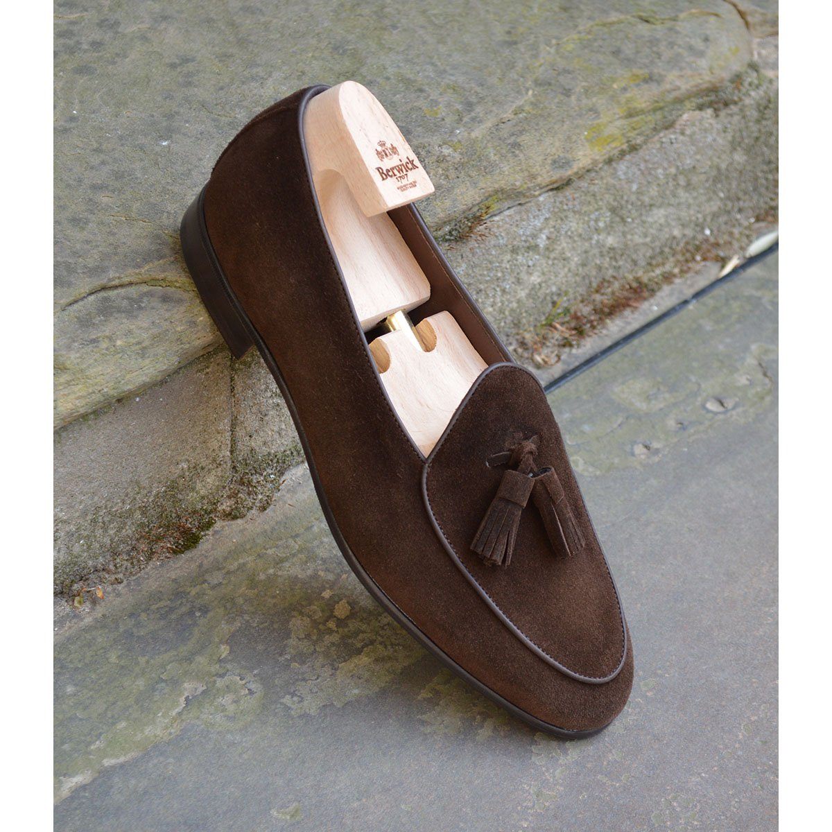 A dark brown suede tassel loafer with a Vegano Moka leather trim, displaying an almond-shaped toe and a fine ridged rubber sole.