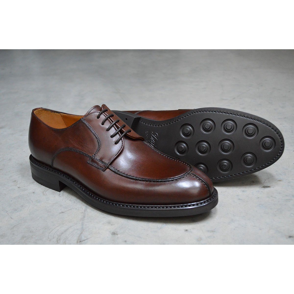 A pair of dark brown Berwick 1707 Split Toe Derby shoes with laces, set against a polished concrete background.