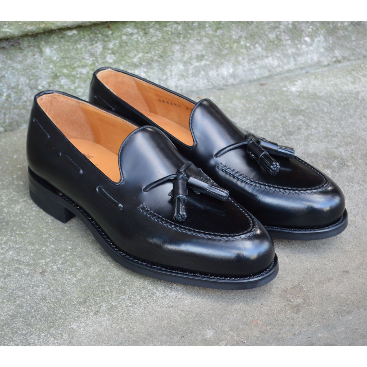A black tasselled slip-on shoe with a polished, smooth upper and a classic round toe. The shoe is set against a natural stone steps background.