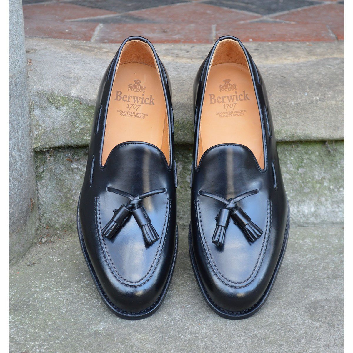 A black tasselled slip-on shoe with a polished, smooth upper and a classic round toe. The shoe is set against a natural stone steps background.