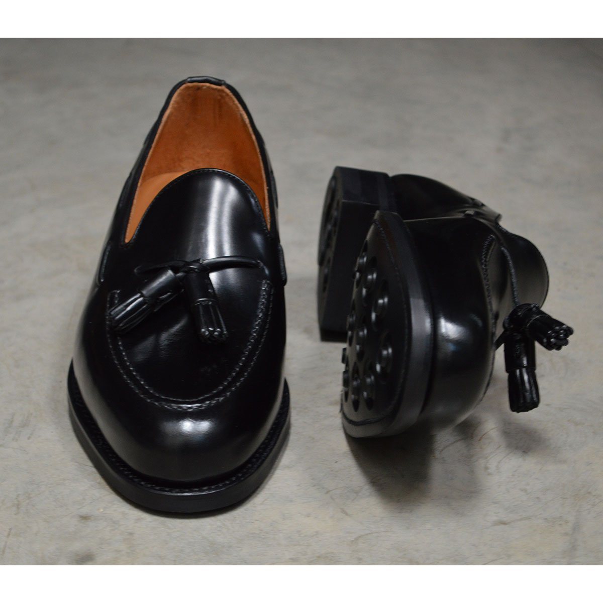 A black tasselled slip-on shoe with a polished, smooth upper and a classic round toe. The shoe is set against a concrete floor background.