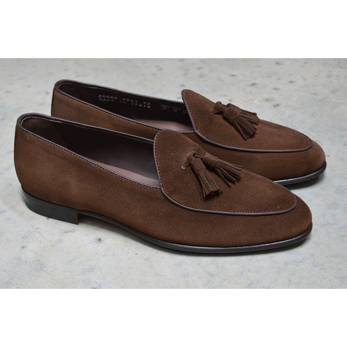 A dark brown suede tassel loafer with a Vegano Moka leather trim, displaying an almond-shaped toe and a fine ridged rubber sole.