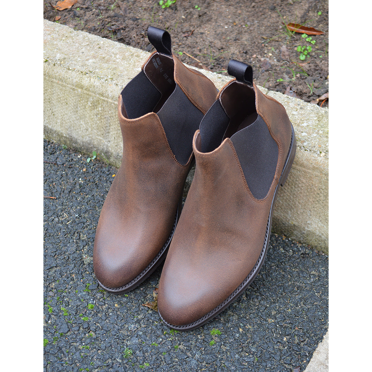 A dark brown Chelsea boot with a waxy leather finish and a contrasting black elastic side panel. The boot has a low heel and a small pull tab at the rear.