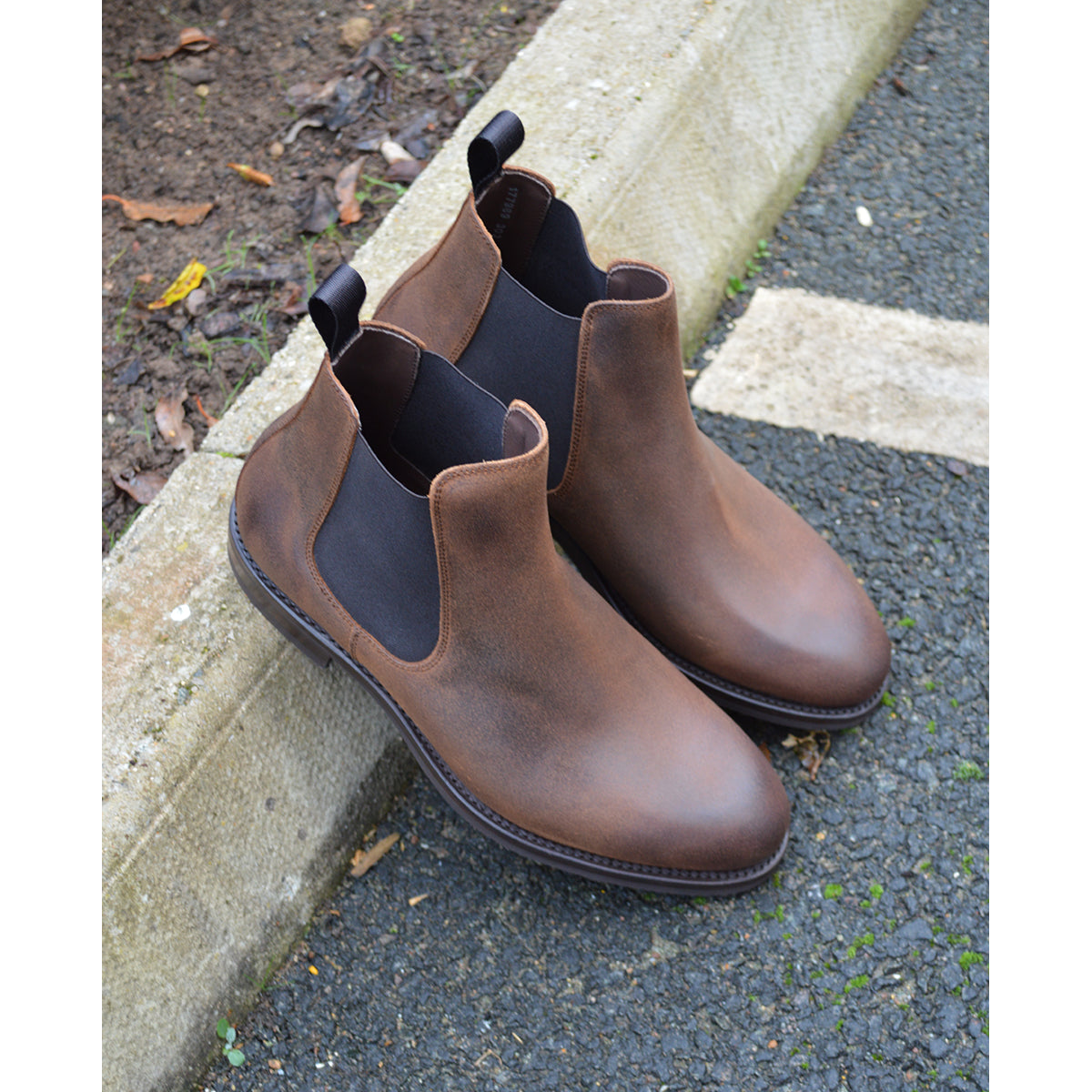 A dark brown Chelsea boot with a waxy leather finish and a contrasting black elastic side panel. The boot has a low heel and a small pull tab at the rear.