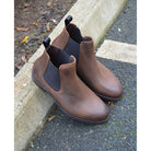 A dark brown Chelsea boot with a waxy leather finish and a contrasting black elastic side panel. The boot has a low heel and a small pull tab at the rear.