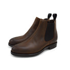 A dark brown Chelsea boot with a waxy leather finish and a contrasting black elastic side panel. The boot has a low heel and a small pull tab at the rear.