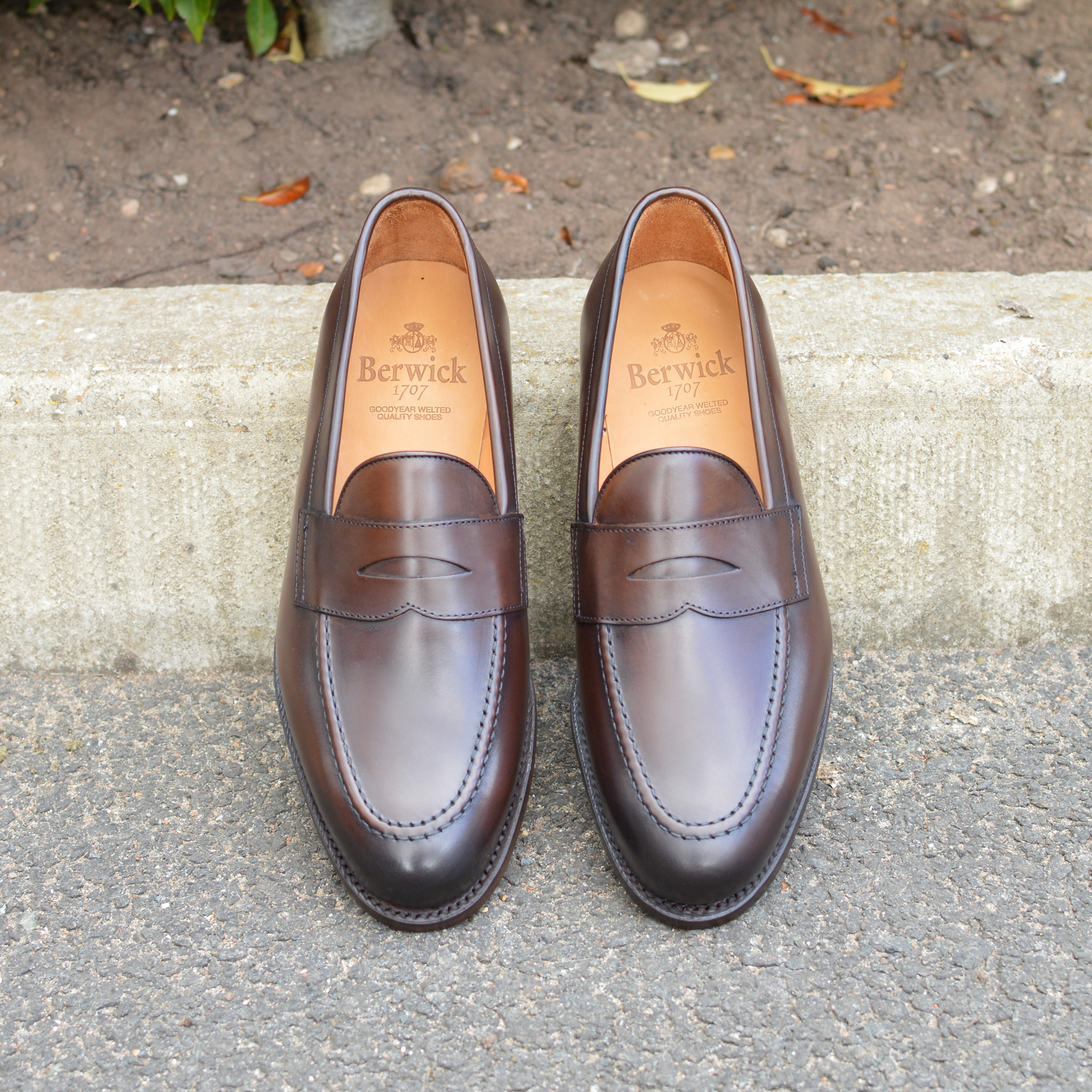 A dark brown Berwick 1707 Penny Loafer with a closed toe and loafers style heel, displayed on a concrete curb background.