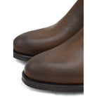 A dark brown Chelsea boot with a waxy leather finish and a contrasting black elastic side panel. The boot has a low heel and a small pull tab at the rear.