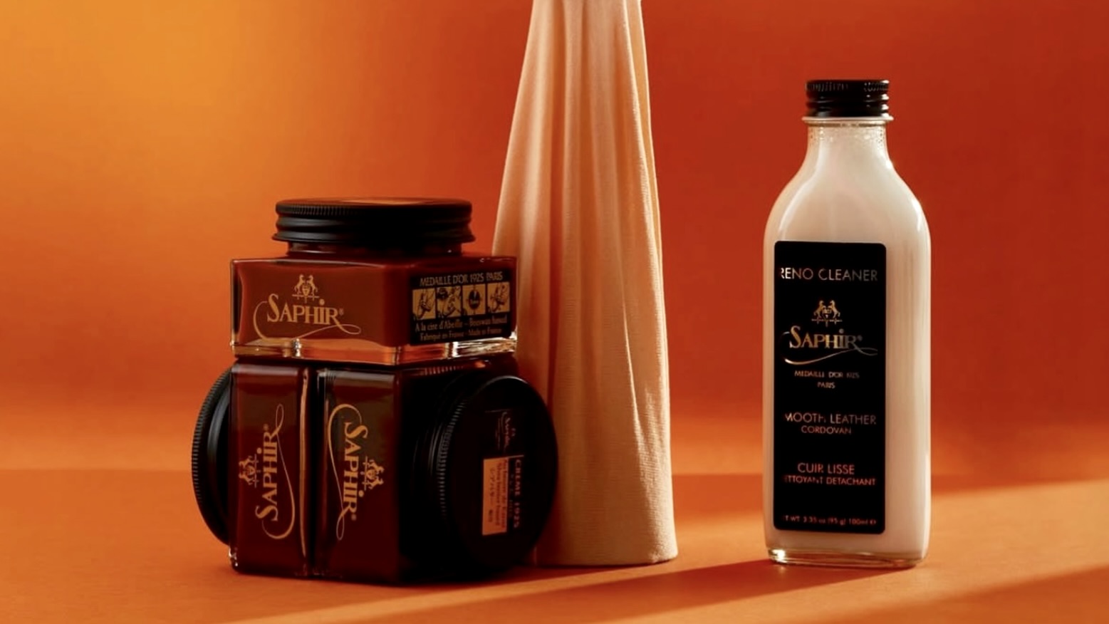 Collection of Saphir Medaille D'Or shoecare products including a bottle of Reno Cleaner and jars of brown Pommadier cream polish on an orange background with a cotton cloth
