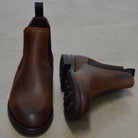 A dark brown Chelsea boot with a waxy leather finish and a contrasting black elastic side panel. The boot has a low heel and a small pull tab at the rear.