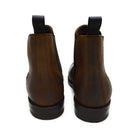 A dark brown Chelsea boot with a waxy leather finish and a contrasting black elastic side panel. The boot has a low heel and a small pull tab at the rear.