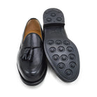 A black deerskin slip-on loafer with a tassel detail on the vamp, featuring a classic round toe and a low heel.
