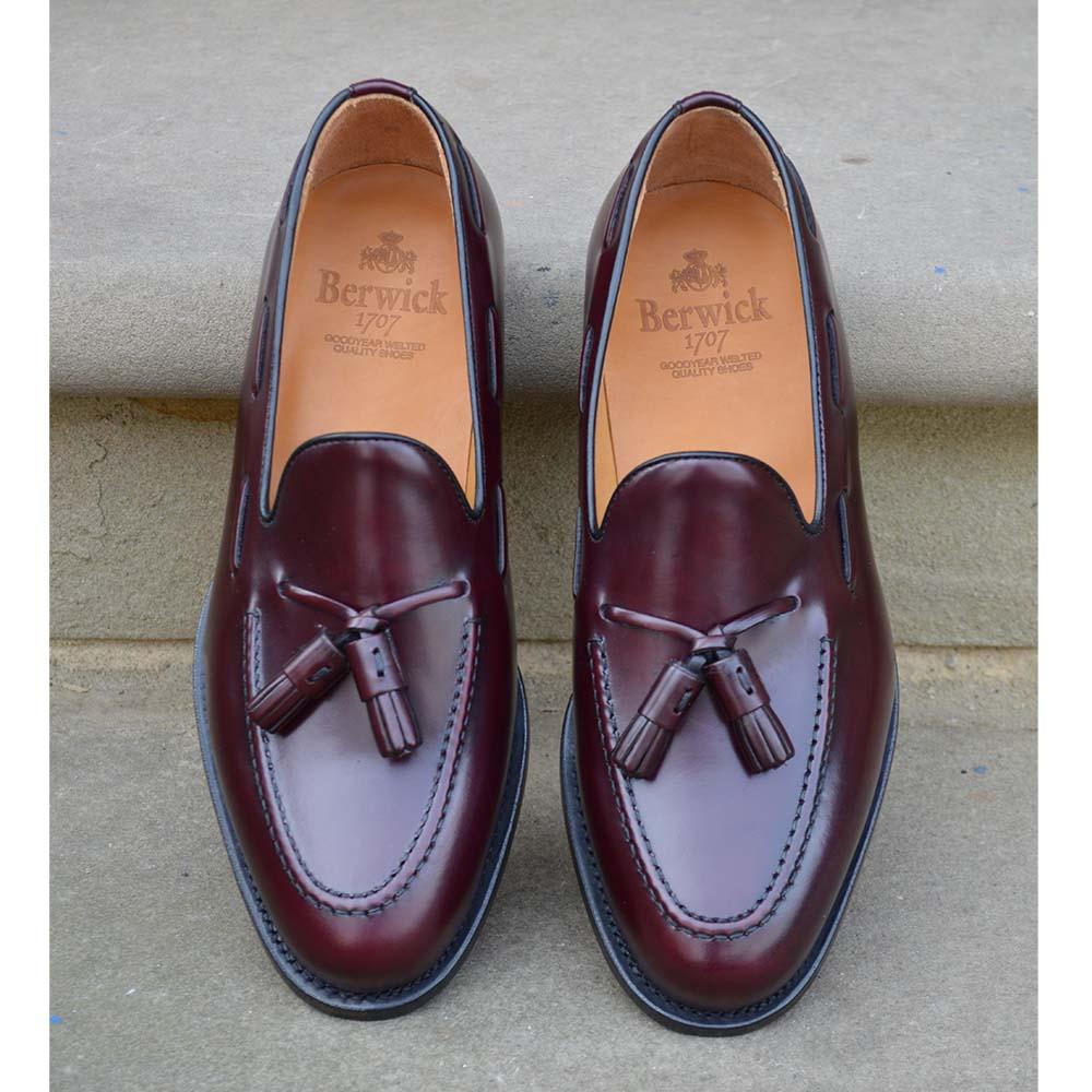 A Berwick 1707 burgundy hi shine leather slip-on shoe with a tassel detail on the vamp, set against a sandstone  background.