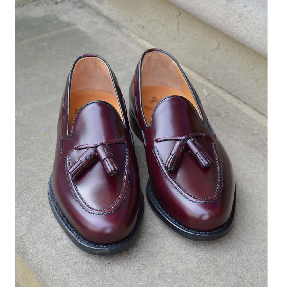 A Berwick 1707 burgundy hi shine leather slip-on shoe with a tassel detail on the vamp, set against a sandstone background.