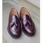 A Berwick 1707 burgundy hi shine leather slip-on shoe with a tassel detail on the vamp, set against a sandstone background.