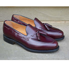 A Berwick 1707 burgundy hi shine leather slip-on shoe with a tassel detail on the vamp, set against a sandstone background.