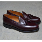 A Berwick 1707 burgundy hi shine leather slip-on shoe with a tassel detail on the vamp, set against a  polished concrete background.