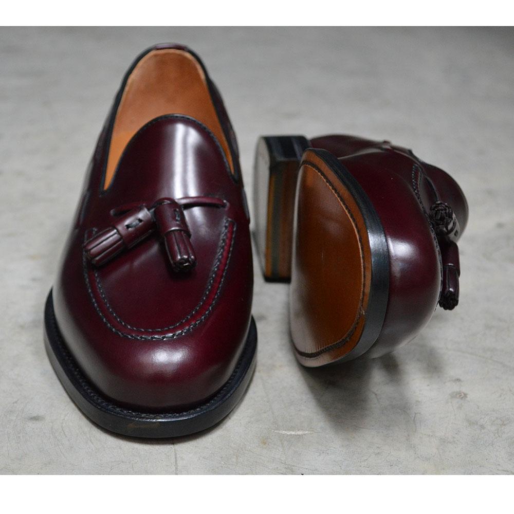A Berwick 1707 burgundy hi shine leather slip-on shoe with a tassel detail on the vamp, set against a polished concrete background.