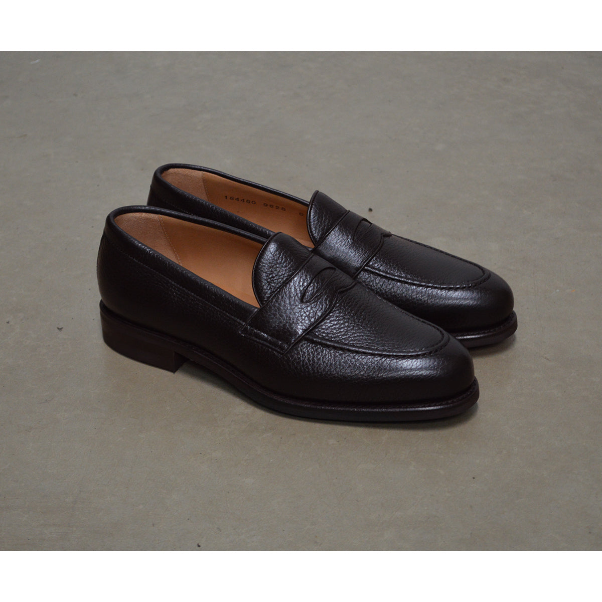 A dark brown deerskin penny loafer with a velvety soft texture and a classic round toe shape on a polished concrete background with Dainite Studded Rubber Sole.