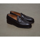 A dark brown deerskin penny loafer with a velvety soft texture and a classic round toe shape on a polished concrete background with Dainite Studded Rubber Sole.