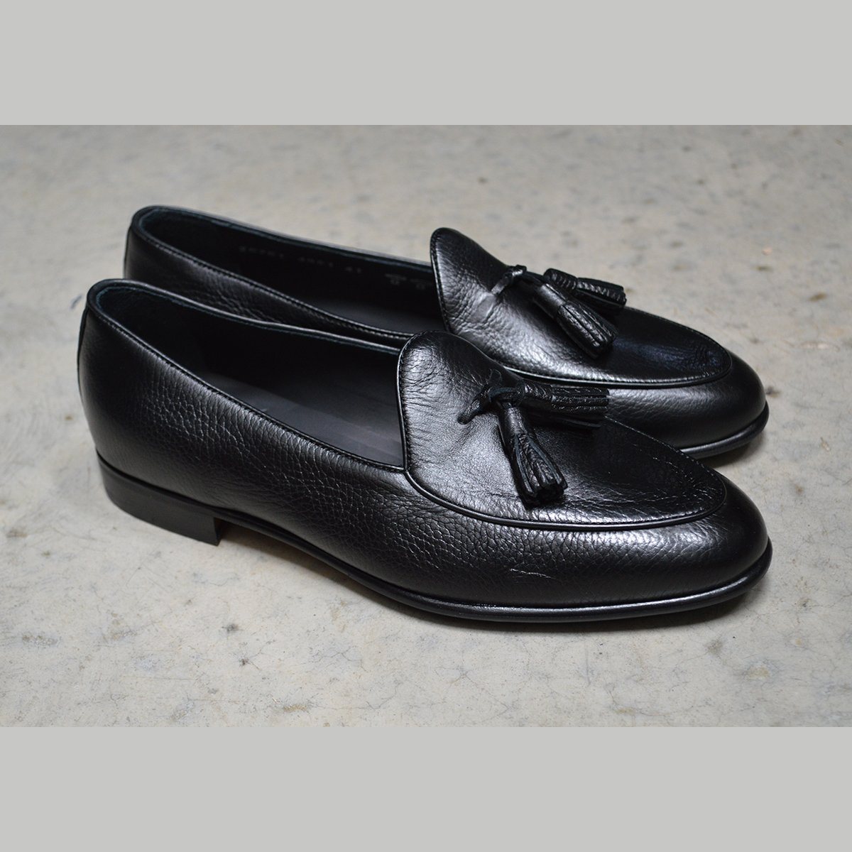 A black deerskin leather tassel Belgian loafer with an almond-shaped toe and a subtle tread sole, displayed against a polished concrete background.