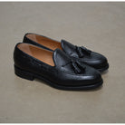 A black deerskin slip-on loafer with a tassel detail on the vamp, featuring a classic round toe and a low heel.