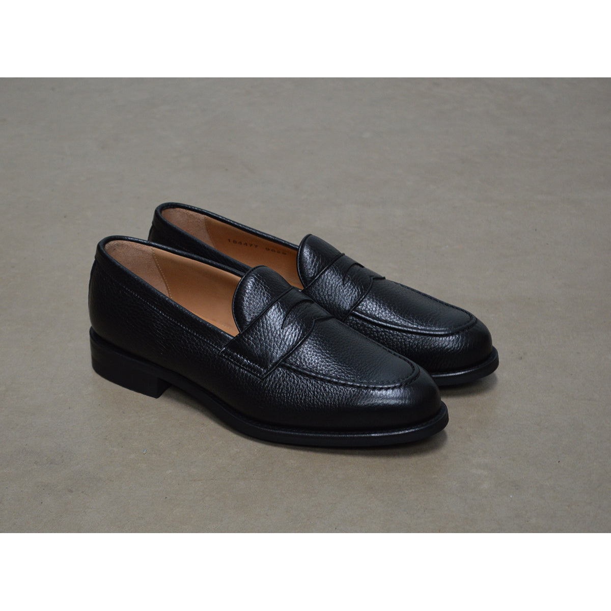 A black deerskin Penny loafer with a velvety soft texture and a classic round toe & Dainite sole, set against a polished concrete floor background.