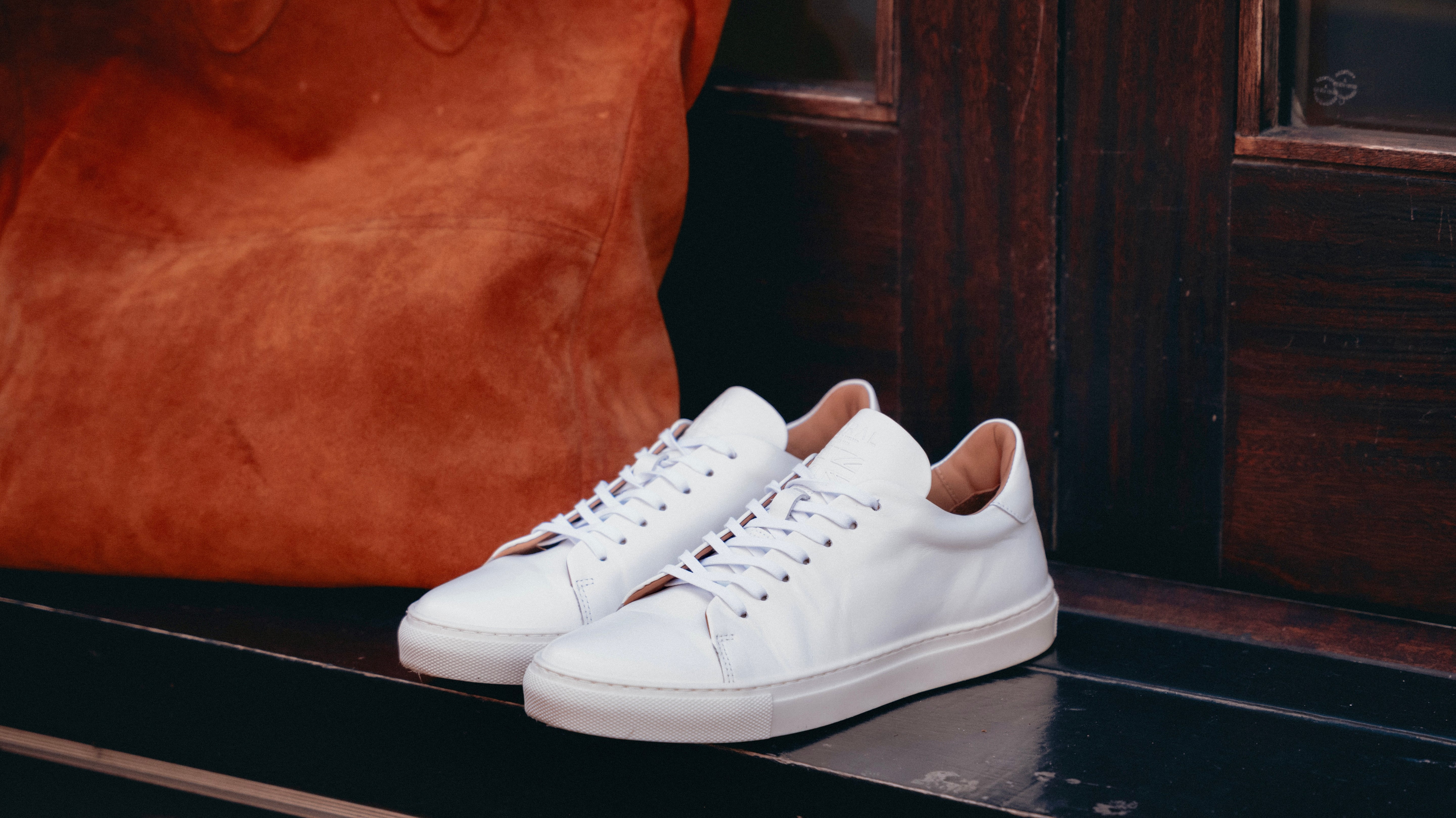 Goral Mellor II Nappa White sneakers on a black surface, wooden doors behind with a rich brown leather tote bag in the background