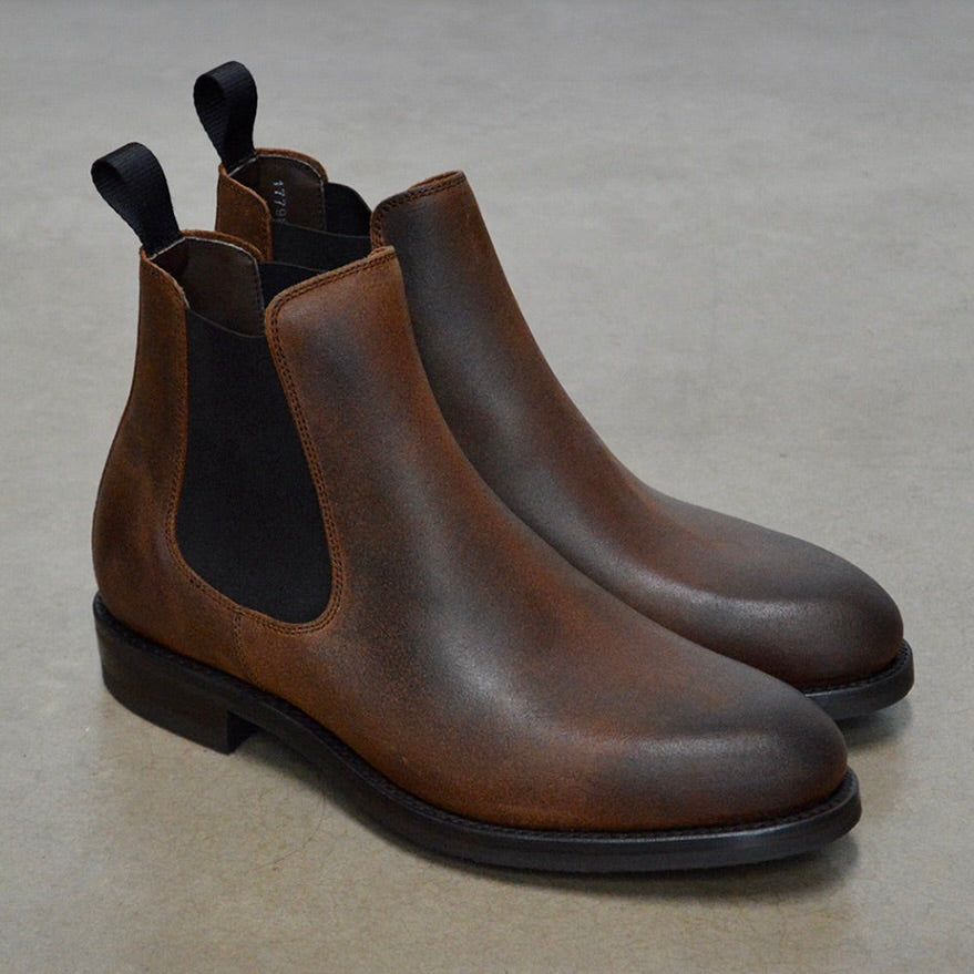 A dark brown Chelsea boot with a waxy leather finish and a contrasting black elastic side panel. The boot has a low heel and a small pull tab at the rear.