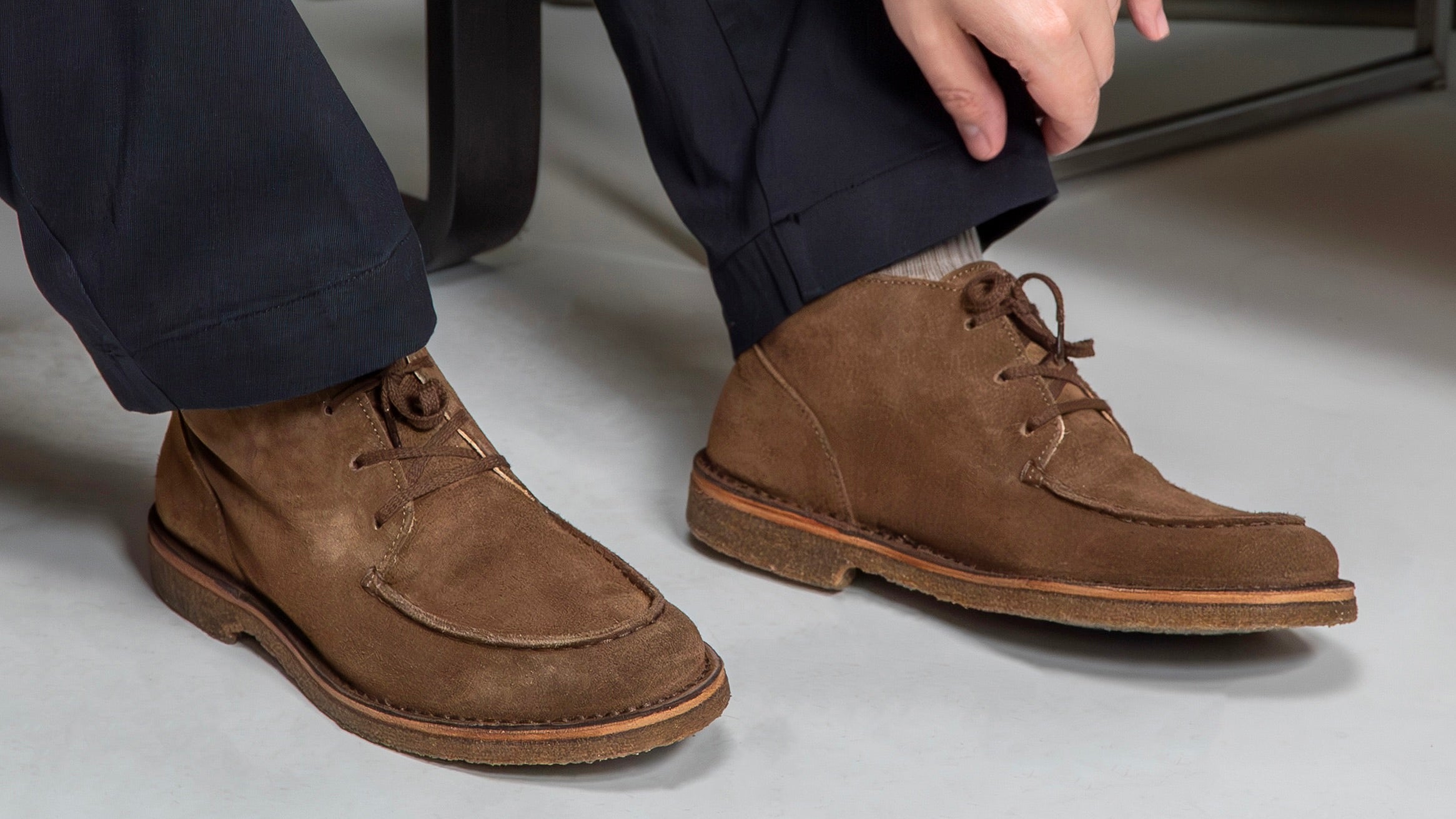 Astorflex Dukeflex apron derby boots in rich Dark Khaki brown suede with crepe rubber soles worn by a smart gentleman sitting on a scandi chair on the grey floor