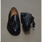 A black deerskin slip-on loafer with a tassel detail on the vamp, featuring a classic round toe and a low heel.