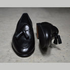 A black deerskin leather tassel Belgian loafer with an almond-shaped toe and a subtle tread sole, displayed against a polished concrete background.