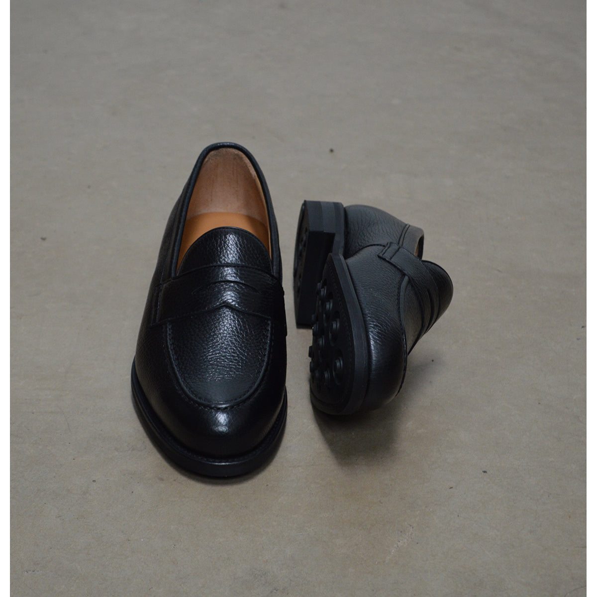 A black deerskin Penny loafer with a velvety soft texture and a classic round toe & Dainite sole, set against a polished concrete floor background.