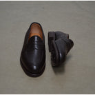 A dark brown deerskin penny loafer with a velvety soft texture and a classic round toe shape on a polished concrete background with Dainite Studded Rubber Sole.