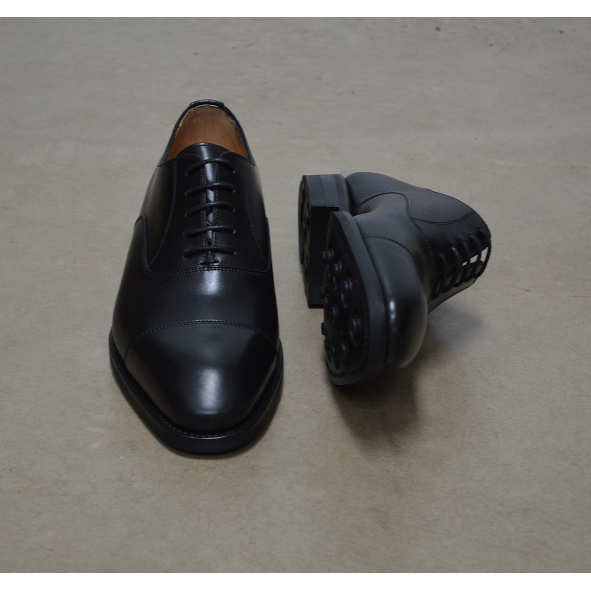 A Berwick 1707 black leather Oxford shoe with a chisel toe and lace-up closure with a Dainite rubber studded sole, on a polished concrete background.